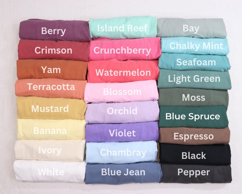 May include: A display of folded t-shirts in various colors, including Berry, Crimson, and Yam. Each shirt is labeled with its color name, such as "Island Reef," "Crunchberry," and "Bay." The shirts are stacked neatly, showcasing a range of hues.