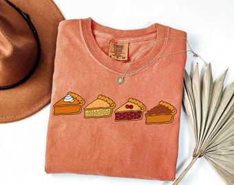 Op de afbeelding: Een perzikkleurig T-shirt met een afbeelding van vier verschillende taartpunten. De punten zijn pompoen, appel, kers en pecan. De tekst "COMFORT COLORS" staat op een label dat aan het shirt is bevestigd.