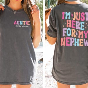 May include: A dark gray t-shirt with a colorful graphic that says "I'm Just Here For My Nephew" on the back and "AUNTY Barbara" on the front.