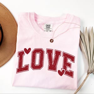 a pink t-shirt with the word "LOVE" printed on it, along with two red heart-shaped symbols. The t-shirt is laid out on a white surface, and there is a brown hat and a fan nearby.