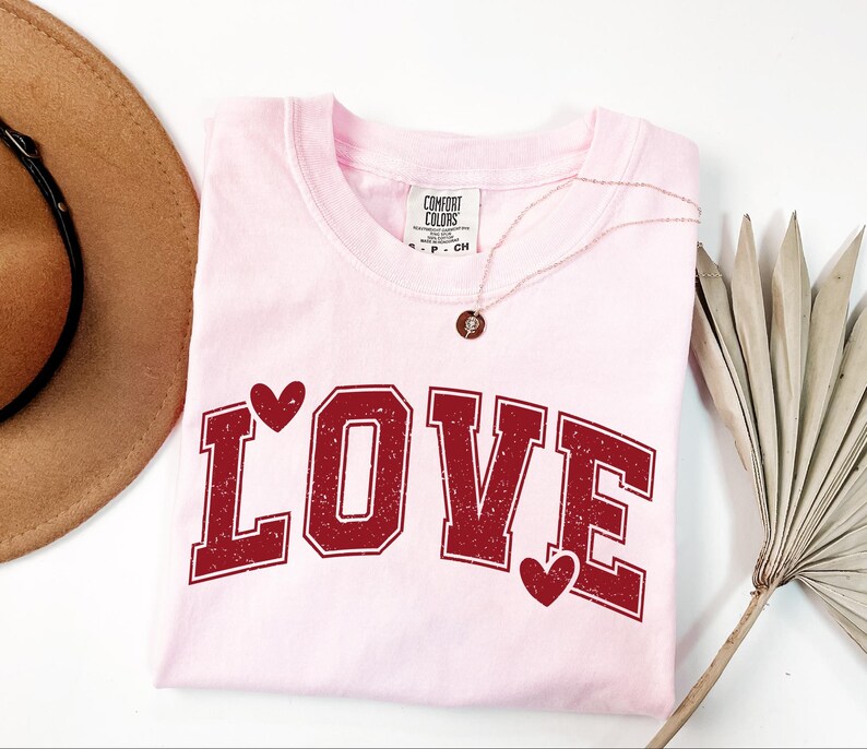 a pink t-shirt with the word "LOVE" printed on it, along with two red heart-shaped symbols. The t-shirt is laid out on a white surface, and there is a brown hat and a fan nearby.