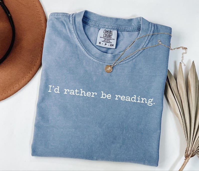 May include: A blue t-shirt with the phrase "I'd rather be reading" in white lettering. A gold necklace with a pendant rests on the shirt. The shirt is folded and has a tag that says "Comfort Colors".