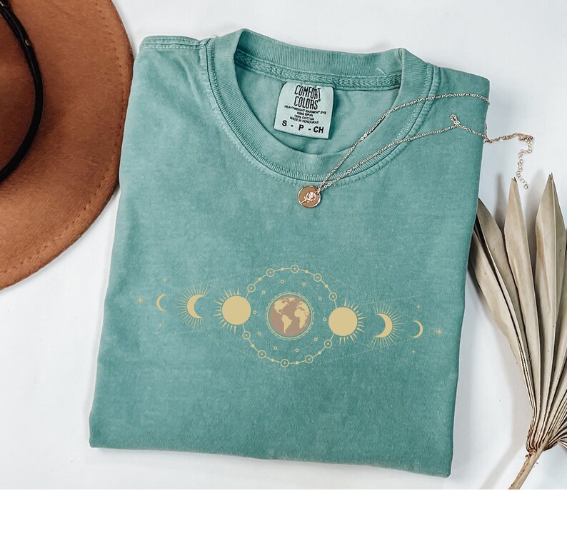 K&ouml;nnte beinhalten: Ein blaues T-Shirt mit einem Grafikdruck eines himmlischen Designs mit Erde, Sonne und Mond. Das Shirt hat ein Etikett mit der Aufschrift "Comfort Colors" und "S - P - CH".