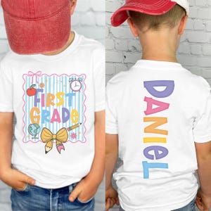 May include: White t-shirt with a colorful "First Grade" design on the front, featuring an apple, globe, pencil, and clock. The back of the shirt displays the name "DANIEL" in vertical, rainbow-colored letters. The shirt is worn by a child.