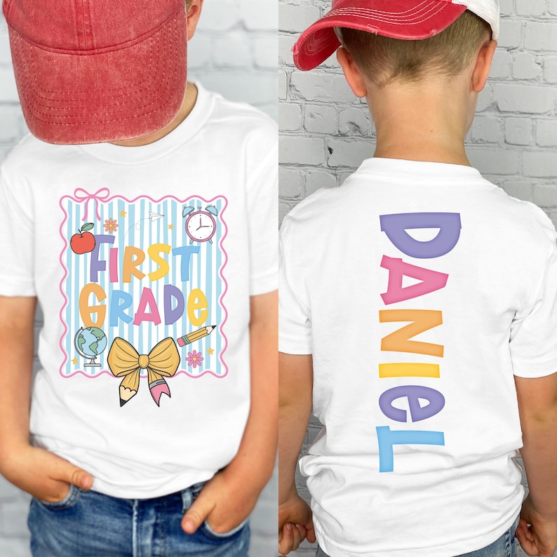 May include: White t-shirt with a colorful "First Grade" design on the front, featuring an apple, globe, pencil, and clock. The back of the shirt displays the name "DANIEL" in vertical, rainbow-colored letters. The shirt is worn by a child.