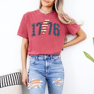 May include: A red t-shirt with a distressed look featuring a white, blue, and red lightning bolt with the number 1776. The lightning bolt is designed to look like the American flag.