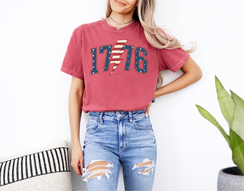May include: A red t-shirt with a distressed look featuring a white, blue, and red lightning bolt with the number 1776. The lightning bolt is designed to look like the American flag.