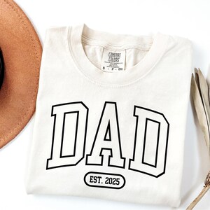 May include: A cream coloured t-shirt with black text that reads "DAD EST. 2025" in a varsity style font.