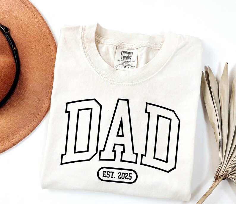 May include: A cream coloured t-shirt with black text that reads "DAD EST. 2025" in a varsity style font.