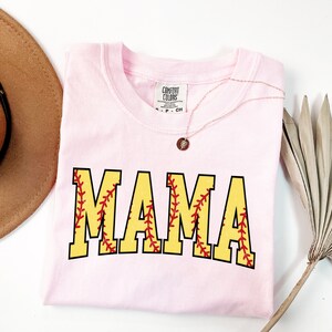 May include: Pink t-shirt with the word "MAMA" in yellow letters with red stitching, resembling a softball. The shirt has a tag that reads "Comfort Colors".