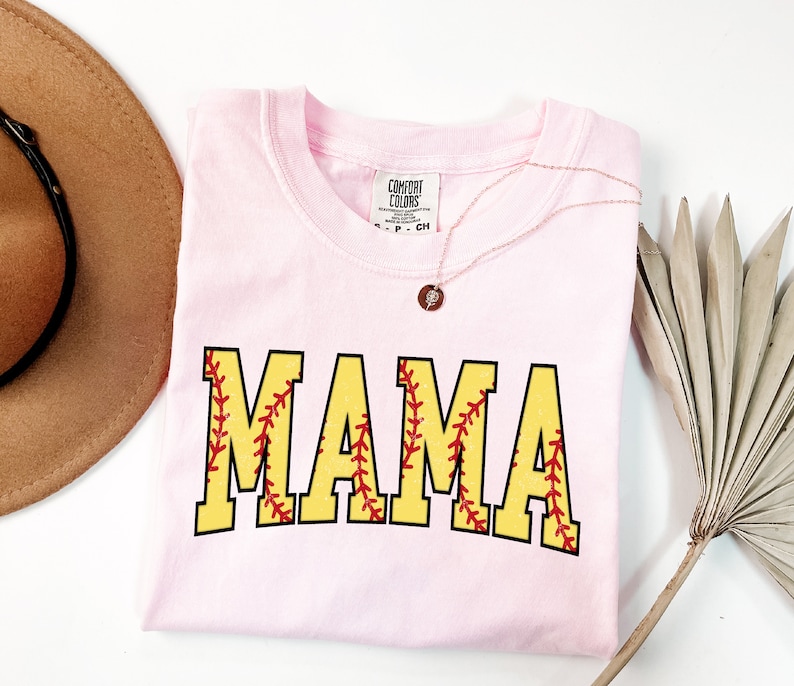 May include: Pink t-shirt with the word "MAMA" in yellow letters with red stitching, resembling a softball. The shirt has a tag that reads "Comfort Colors".