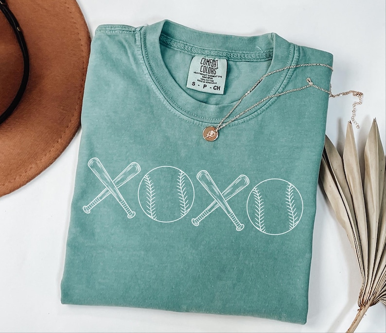 May include: A teal green t-shirt featuring a white graphic of two baseball bats crossed over each other with a baseball on either side of the bats. The graphic says "XOXO" with the baseballs forming the "O". The shirt has a tag that says "Comfort Colors" with the sizes S-P-CH.