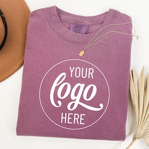 May include: A mauve coloured t-shirt with a white circle that says "YOUR logo HERE" in a cursive font.