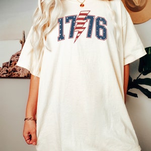 May include: A white oversized t-shirt with a red, white, and blue lightning bolt graphic and the text "1776".