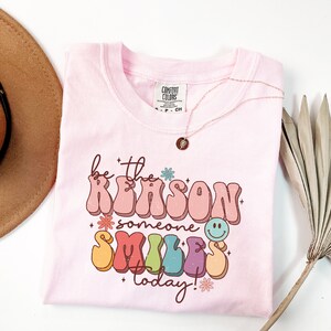 May include: Pink t-shirt with a retro-style graphic that says "be the reason someone smiles today!" The graphic is in rainbow colors with a smiley face and flowers.