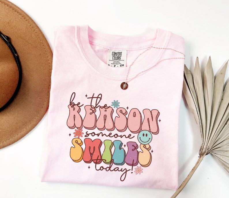 May include: Pink t-shirt with a retro-style graphic that says "be the reason someone smiles today!" The graphic is in rainbow colors with a smiley face and flowers.