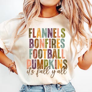 May include: Cream-colored t-shirt with the words "Flannels, Bonfires, Football, Pumpkins, it's fall y'all" in a colorful, stacked design. The shirt is a casual style, perfect for autumn.