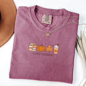 May include: A mauve-coloured t-shirt with a crew neck and short sleeves. The shirt features a graphic of books, a pumpkin, a leaf, and a coffee cup with the words "cozy season" underneath. A gold necklace is draped across the shirt.