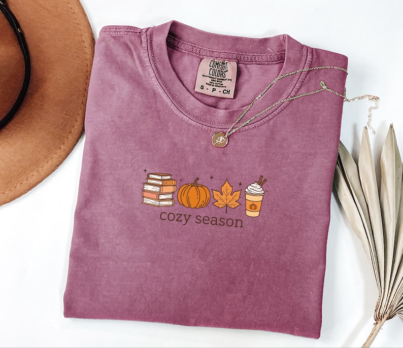 May include: A mauve-coloured t-shirt with a crew neck and short sleeves. The shirt features a graphic of books, a pumpkin, a leaf, and a coffee cup with the words "cozy season" underneath. A gold necklace is draped across the shirt.