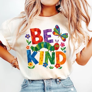 May include: A cream-colored short-sleeve t-shirt featuring a colorful graphic print. The design includes the text BE KIND in large, multi-colored letters, a green caterpillar, a multi-colored butterfly, and various small flowers in red, blue, and purple.