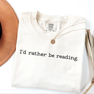 May include: Cream-colored t-shirt with the text "I'd rather be reading" in black. A gold necklace with a pendant rests on the shirt. A brown hat and dried plant are in the background.