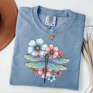 May include: A blue Comfort Colors t-shirt featuring a colorful dragonfly design with flowers. The dragonfly has iridescent wings and is surrounded by pink, blue, and yellow flowers. A gold necklace rests on the shirt.