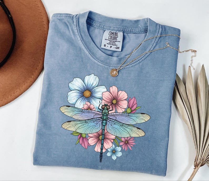 May include: A blue Comfort Colors t-shirt featuring a colorful dragonfly design with flowers. The dragonfly has iridescent wings and is surrounded by pink, blue, and yellow flowers. A gold necklace rests on the shirt.