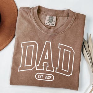 May include: A brown t-shirt with a white "DAD" graphic in a varsity style font. The shirt also has a white circle with the text "EST. 2025" below the graphic.