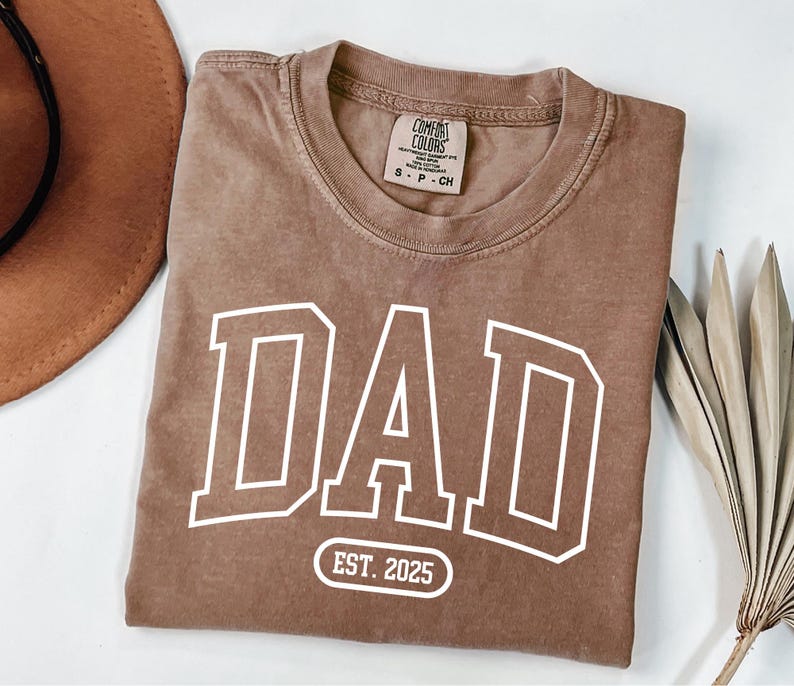 May include: A brown t-shirt with a white "DAD" graphic in a varsity style font. The shirt also has a white circle with the text "EST. 2025" below the graphic.