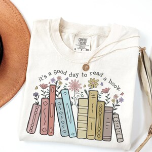 K&ouml;nnte beinhalten: Cremefarbenes T-Shirt mit dem Text "it's a good day to read a book" &uuml;ber einer Reihe von illustrierten B&uuml;chern und Blumen. Eine goldene Halskette mit Anh&auml;nger liegt auf dem Shirt. Das Shirt ist von der Marke Comfort Colors.