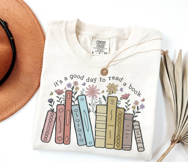 K&ouml;nnte beinhalten: Cremefarbenes T-Shirt mit dem Text "it's a good day to read a book" &uuml;ber einer Reihe von illustrierten B&uuml;chern und Blumen. Eine goldene Halskette mit Anh&auml;nger liegt auf dem Shirt. Das Shirt ist von der Marke Comfort Colors.