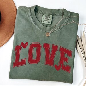 a green t-shirt with the word "LOVE" printed on it, along with red heart shapes. The t-shirt is placed on a white surface, and there is a brown hat and some dried leaves nearby.