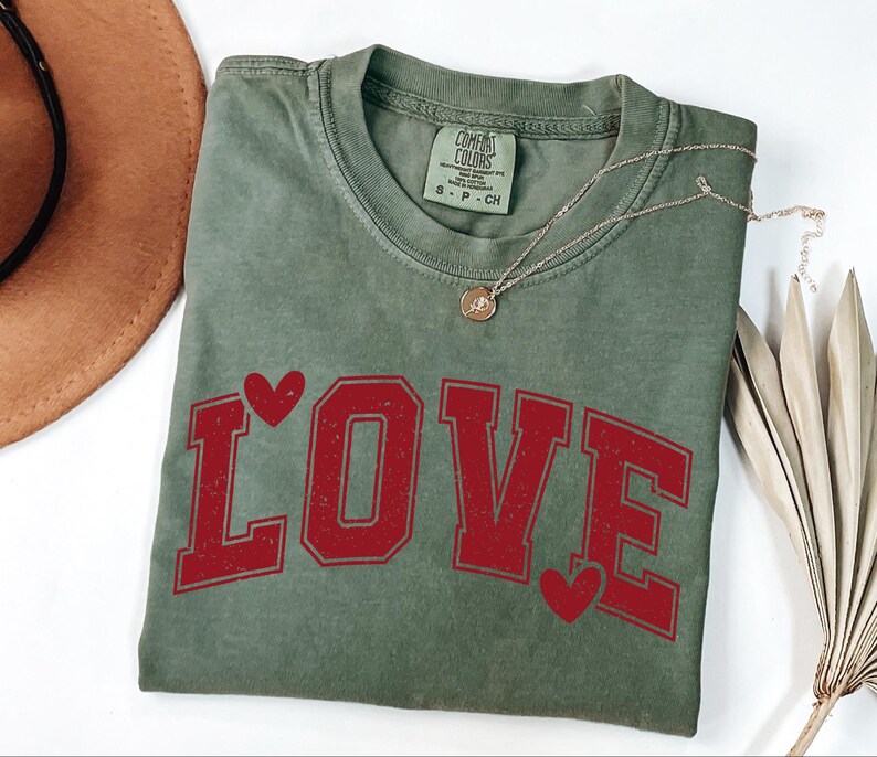 a green t-shirt with the word "LOVE" printed on it, along with red heart shapes. The t-shirt is placed on a white surface, and there is a brown hat and some dried leaves nearby.