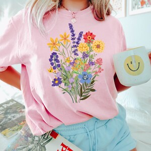 May include: A pink t-shirt with a colorful floral bouquet graphic print. The shirt is worn by a person with blonde hair and blue shorts. The person is holding a light blue mug with a yellow smiley face.