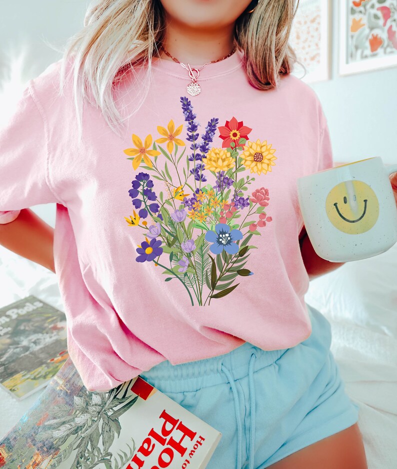 May include: A pink t-shirt with a colorful floral bouquet graphic print. The shirt is worn by a person with blonde hair and blue shorts. The person is holding a light blue mug with a yellow smiley face.