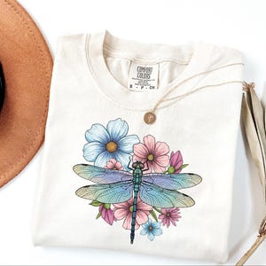 May include: Cream-colored t-shirt featuring a colorful dragonfly design with blue, pink, and green hues, surrounded by flowers. A gold necklace and a brown hat are visible in the background. The shirt has the Comfort Colors brand tag.