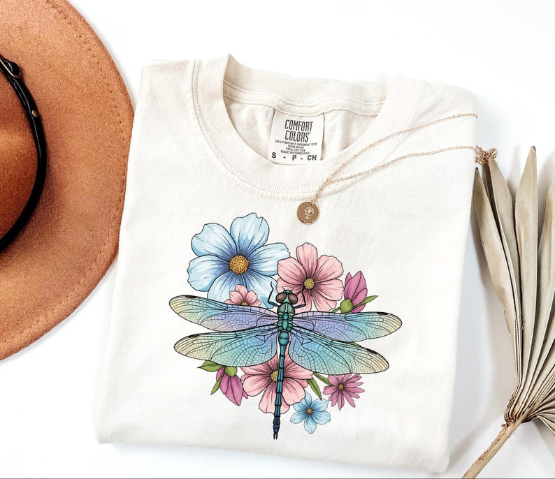 May include: Cream-colored t-shirt featuring a colorful dragonfly design with blue, pink, and green hues, surrounded by flowers. A gold necklace and a brown hat are visible in the background. The shirt has the Comfort Colors brand tag.