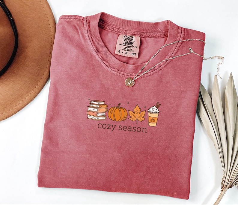 May include: A maroon Comfort Colors t-shirt featuring a fall-themed graphic. The graphic includes a stack of books, a pumpkin, a leaf, and a coffee cup, with the words "cozy season" below. A silver necklace with a pendant is draped on the shirt.