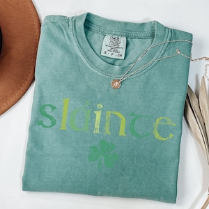 May include: A teal green t-shirt with the word "slainte" in green, yellow, and white lettering. A green four-leaf clover is below the word.