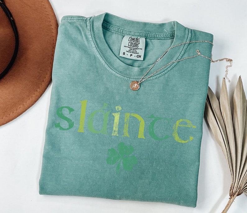 May include: A teal green t-shirt with the word "slainte" in green, yellow, and white lettering. A green four-leaf clover is below the word.