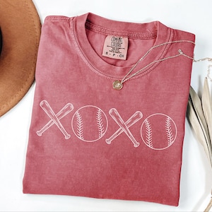 May include: A red t-shirt featuring a white graphic of two baseball bats crossed over each other with a baseball on either side of the bats. The graphic says "XOXO" with the baseballs forming the "O". The shirt has a distressed look and is made of a soft, comfortable material.