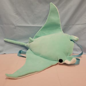 May include: A plush stingray backpack in a soft mint green color with a white underside. The bag features a long tail, two black circular eyes, and light blue adjustable nylon straps attached to the back for carrying as a functional accessory.