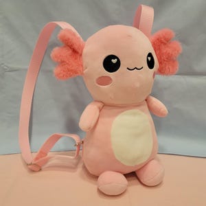 Pode incluir: Uma mochila de pelúcia em formato de axolote rosa claro com barriga creme. A criatura possui guelras rosas felpudas, olhos bordados em preto com reflexos brancos em formato de coração, um pequeno sorriso e bochechas rosas. Alças ajustáveis.
