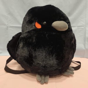 May include: A plush backpack shaped like a round black bird. The bird has soft black fur, a gray beak, gray feet, and a small orange patch near its black eye. Two black adjustable straps are attached to the back of the plush bird figure.