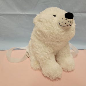 Puede incluir: Una mochila de peluche con forma de oso polar blanco, hecha de piel sintética suave y peluda, con una nariz negra bordada y una boca cosida. Incluye correas de tela blanca ajustables con herrajes de metal plateado para llevarla puesta.