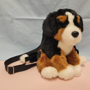 Pode incluir: Uma mochila de pelúcia em forma de boiadeiro bernês com pelo macio preto, castanho e branco. A bolsa possui uma alça de nylon preta ajustável com um regulador de metal prateado, um nariz de plástico preto e patas brancas.