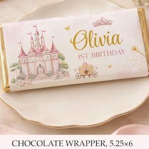 以下が含まれることがあります： 5.25 x 6インチの長方形のチョコレートバー用ラッパー。ピンクの城のイラスト、金色の馬車、ティアラ、魔法の杖が描かれています。テキストは金とピンクのスクリプト体とブロック体で「Olivia 1ST BIRTHDAY」と記されています。