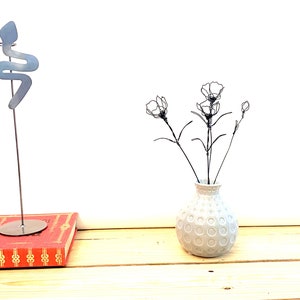 May include: A white ceramic vase with a textured surface, featuring a circular pattern, holds a bouquet of black wire flowers. The vase is sitting on a wooden surface.