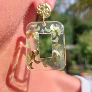May include: A rectangular clear resin earring featuring embedded gold-colored crescent moon shapes and a small white star. The earring hangs from a circular gold-colored stud with a textured, hammered finish, creating a reflective appearance.