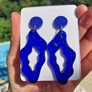 Puede incluir: Un par de pendientes azul vibrante con una forma abstracta única. Cada pendiente presenta una parte superior circular y un diseño abierto y fluido con detalles de purpurina. Los pendientes se muestran en una tarjeta blanca.